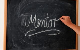 Main écrivant le mot "Mentor" sur un tableau noir